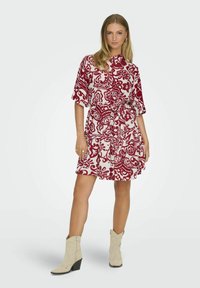 Abito a fiori rosso e bianco con maniche corte, vita con allaccio e orlo con ruches. Il modello indossa stivali da cowboy beige.