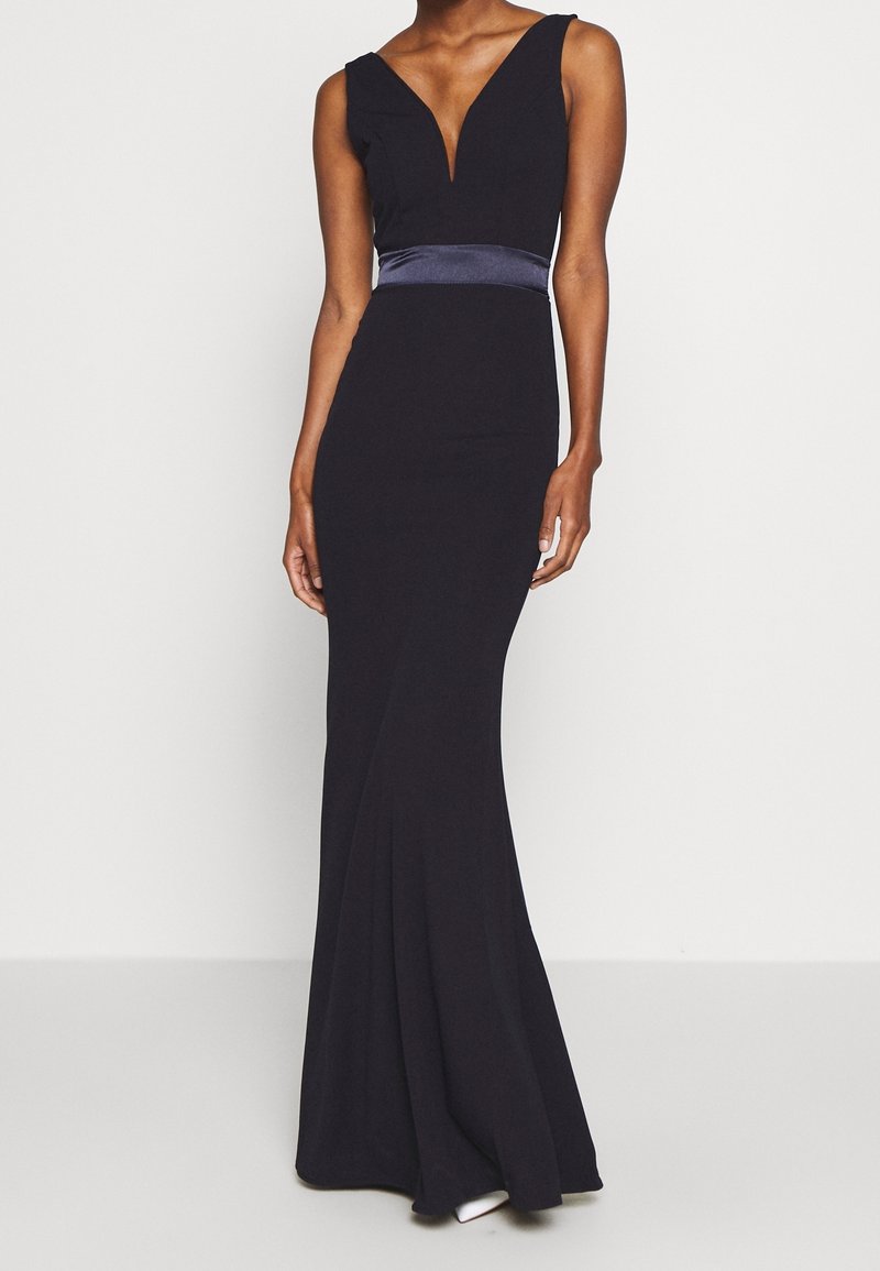 Robe de soirée noire sans manches avec un profond décolleté en V, taille ajustée avec une ceinture sombre brillante et une jupe évasée longue jusqu'au sol portée par une personne debout.