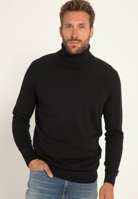 Schwarzer Rollkragenpullover mit langen Ärmeln, strukturierter Stoff, gerippter Besatz am Kragen und Saum, taillierte Form, getragen mit blauen Jeans, Hände in den Taschen.