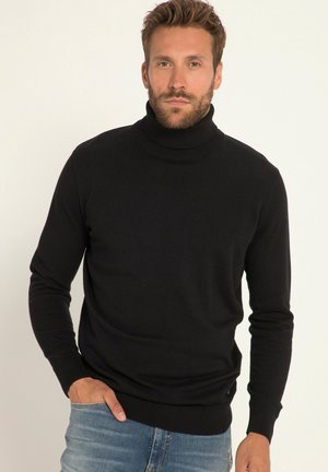 Suéter negro de cuello alto con mangas largas, tejido texturizado, ribetes en el cuello y el dobladillo, forma ajustada, llevado con jeans azules y manos en los bolsillos.
