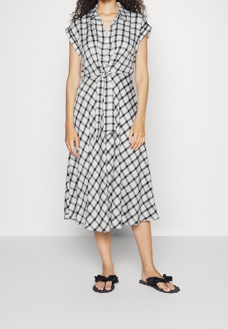 Černobílé kostkované midi šaty se zavázaným pasem, krátkými rukávy a volným lemem. Nosič má na sobě černé žabky s volánky.