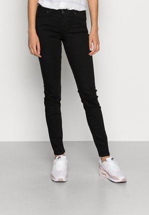 Jeans skinny negros fabricados en denim con una textura suave. Combinados con zapatillas Nike blancas y rosas, de pie sobre un suelo gris.