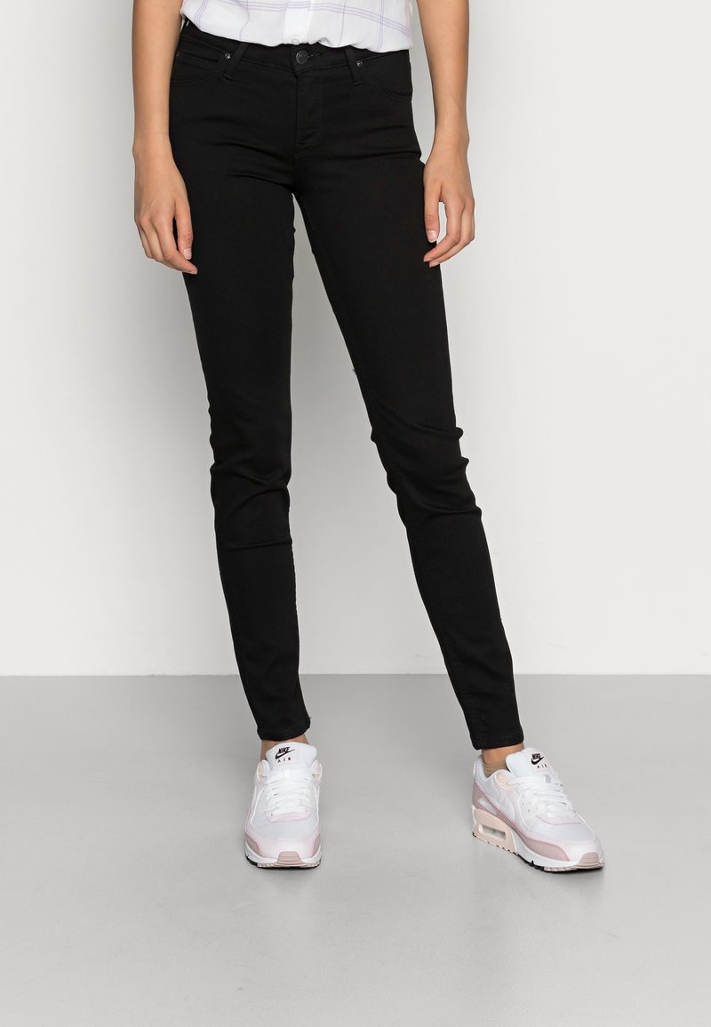 Jeans skinny negros fabricados en denim con una textura suave. Combinados con zapatillas Nike blancas y rosas, de pie sobre un suelo gris.