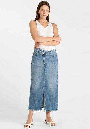 Femme debout, bras croisés, portant un haut blanc sans manches, une longue jupe en jean bleue fendue à l'avant, et des sandales blanches à talons.