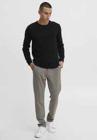 Pull en tricot noir, pantalon gris fuselé, baskets blanches. Texture lisse, poignets côtelés, branding subtil sur le pantalon. Debout avec les mains dans les poches.