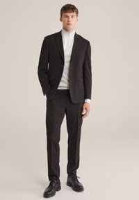 Uskožen slim-fit crni odijelo s rezanim kragnom, upareno s bijelim duksericom na zip. Teksturirani materijal i jednostavne crne cipele za odijelo upotpunjuju izgled.