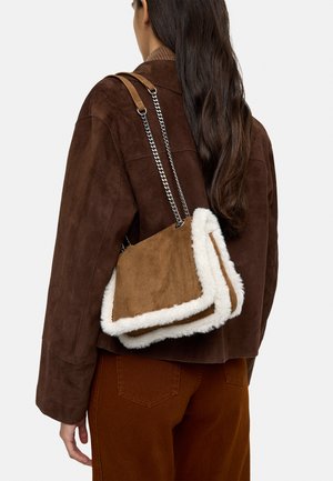 Kobieta w brązowej zamszowej kurtce i spodniach, niosąca na ramieniu brązową zamszową torbę z białym futrzanym obszyciem i srebrnymi łańcuszkowymi ramiączkami.