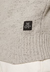 Pull en maille gris clair avec une texture côtelée et une étiquette noire avec des lettres blanches. Le tissu semble doux et confortable.