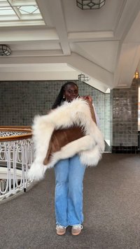 Kunstpelzjacke mit braunem Wildlederkörper und weißem, flauschigem Besatz. Model trägt hellblaue Denim-Flares und beige Sneaker auf einem teppichbedeckten Boden.