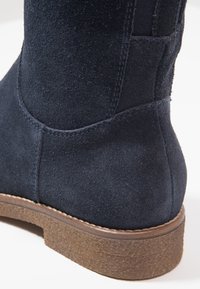 Gros plan sur une botte cheville en daim bleu foncé avec une semelle marron texturée et des coutures visibles sur fond blanc.