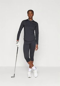 Svart långärmad träningströja med halvdragkedja, tillsammans med svarta capribyxor. Tillbehör inkluderar en vit golfhandke och en golfklubba.