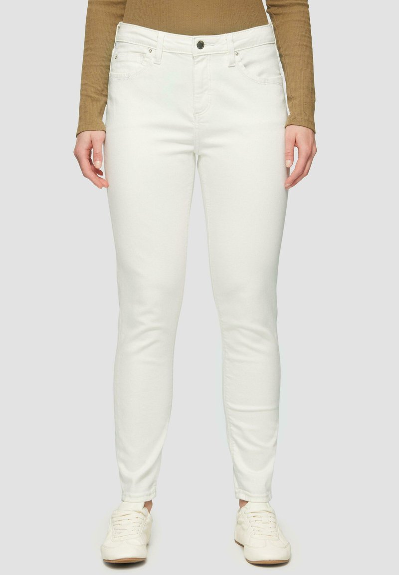 Jeans skinny bianchi con una texture liscia, cinque tasche e chiusura con bottone. Abbinati a sneakers bianche e a una camicia a maniche lunghe marrone.