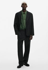 Fekete zakó kontrasztos varrással, zöld gombos ing fölött viselve. Laza, bőszárú nadrág és fekete cipő egészíti ki az öltözéket.