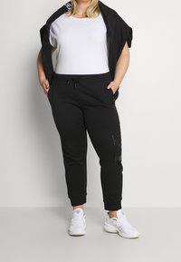 Schwarze Jogginghosen mit elastischem Bund und Kordelzug, ausgestattet mit Seitentaschen und einem subtilen Logodetail am Bein, kombiniert mit einem weißen T-Shirt.