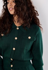 Zielony dzianinowy top z kołnierzem, ozdobiony złotymi guzikami, z dwiema kieszeniami na piersi oraz prążkowanym dołem dla tekstury i dopasowania.