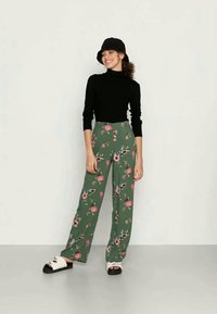 Calças florais verdes de pernas largas, com flores rosas e brancas; combinadas com um suéter preto de gola alta e um chapéu de balde preto. Sandálias brancas.