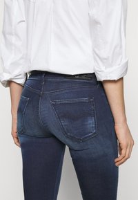 Vaqueros de mezclilla elásticos de color azul oscuro con una cintura de altura media, dos bolsillos traseros con un patrón de costura y un efecto de desvanecimiento sutil.