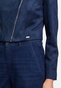 Marineblaue, synthetische Wildlederjacke mit silbernem Reißverschluss und kleinen Logodetails, kombiniert mit Jeans in passendem Farbton.