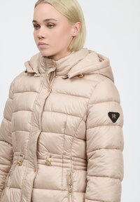 Giubbotto imbottito beige chiaro con cappuccio, texture trapuntata, hardware color oro, tasche laterali e logo sul maniche sinistro.