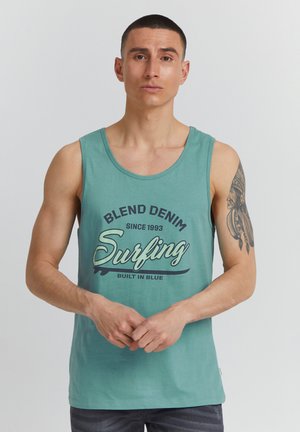 Mężczyzna o krótkich włosach w turkusowej koszulce bez rękawów z napisem "Blend Denim Surfing" oraz tatuażem na prawym ramieniu.