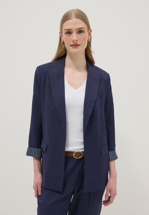 Vrouw draagt een marineblauwe blazer met opgerolde gestreepte manchetten, een wit V-hals topje, marineblauwe broek en een bruine riem met gouden gesp, staand tegen een effen achtergrond.