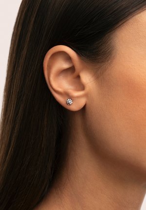 Pendiente de plata con un diseño floral que presenta una gema central transparente. El pendiente se muestra contra un cabello liso y oscuro y una piel tonal.
