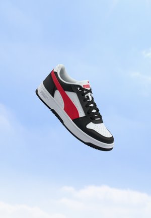 Baskets Puma noires, blanches et rouges avec lacets flottant sur un fond de ciel bleu.