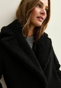 Joven mujer con cabello castaño claro, vestida con un abrigo de lana negra texturizado y un suéter gris, mirando ligeramente hacia arriba y hacia un lado.