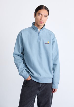 Joven con cabello largo que lleva un sudadero de media cremallera azul claro y pantalones oscuros, de pie con una mano en el bolsillo frente a un fondo blanco.