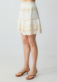 Jupe blanche à volants avec bord dentelé et taille smockée, au design superposé et à la texture douce, complétée par des tongs beige clair.