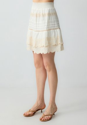 Jupe blanche à volants avec bord dentelé et taille smockée, au design superposé et à la texture douce, complétée par des tongs beige clair.