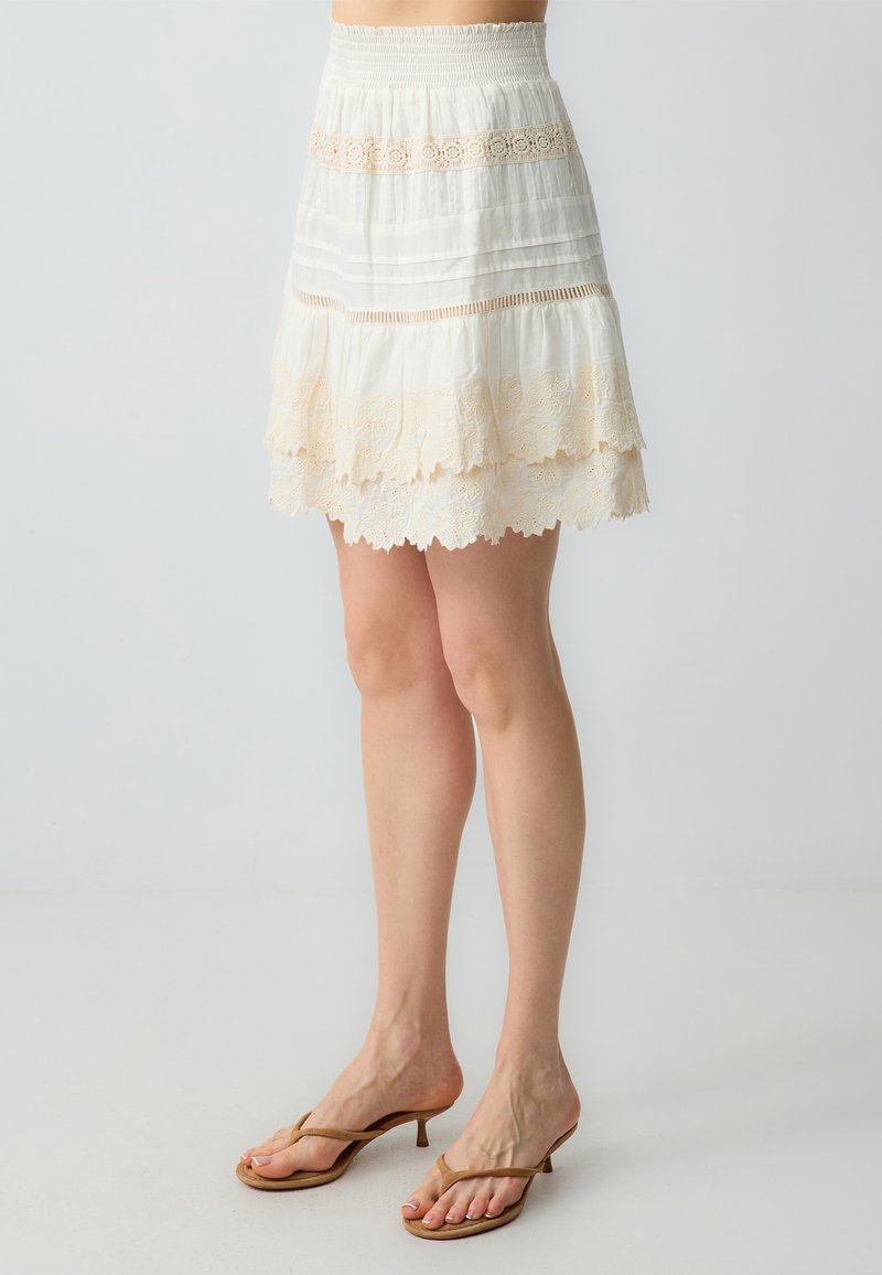 Jupe blanche à volants avec bord dentelé et taille smockée, au design superposé et à la texture douce, complétée par des tongs beige clair.