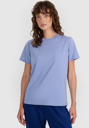 Kobieta o kręconych brązowych włosach, ubrana w jasnofioletowy t-shirt z krótkim rękawem i ciemnoniebieskie spodnie, stojąca na jednolitym tle.