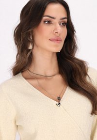 Strick, cremefarbener Wickelsweater mit V-Ausschnitt und strukturiertem Stoff. Verfügt über gestufte Silberhalsketten und ein herzförmiges silbernes Detail.