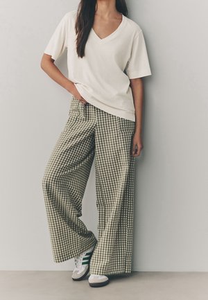 Femme portant un T-shirt blanc ample à col en V, un pantalon large à carreaux et des baskets blanches avec des rayures vertes, debout contre un mur uni.