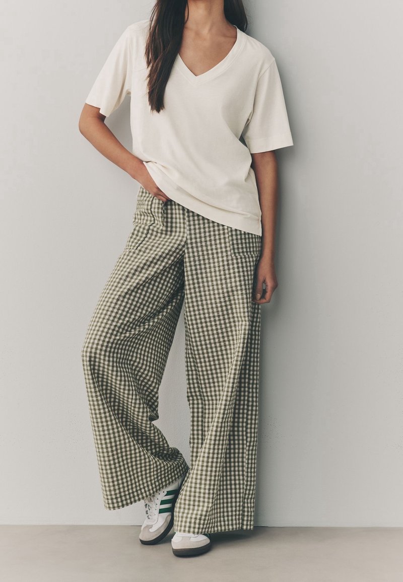 Femme portant un T-shirt blanc ample à col en V, un pantalon large à carreaux et des baskets blanches avec des rayures vertes, debout contre un mur uni.