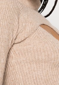 Beiger Strickpullover mit gerippter Textur, hoher Kragen und einem Ausschnitt an einer Schulter. Weiches, warmes Material.