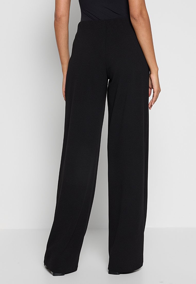 Femme portant un pantalon noir large taille haute et un haut noir ajusté, se tenant devant un fond clair simple.