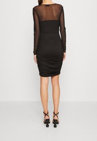 Czarna sukienka z długimi, przezroczystymi rękawami, dopasowanym krojem, marszczeniami na spódnicy oraz gładką fakturą. Czarne sandały na obcasie z okrągłymi akcentami.