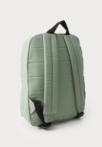 Mochila de tela verde con diseño acolchado, correas ajustables negras y un asa superior. Cuenta con un bolsillo lateral y una construcción robusta.