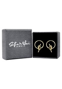 Guldtonade örhängen med cirkulär design, prydda med en knuten detalj. Presenteras i en strukturerad grå låda med svart inredning.