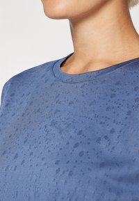 Camicia blu a maniche corte con scollatura tonda, motivo strutturato e finitura in tessuto liscia. Nessun hardware o accenti visibili.