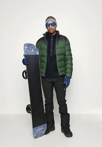 Groene pufferjack, marineblauw shirt met lange mouwen, zwarte broek, blauwe handschoenen en een zonnebril. Houdt een snowboard vast met blauwe accenten.