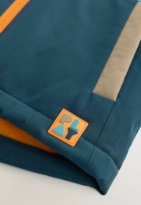 Blauer Stoff mit glatter Textur, ausgestattet mit einem beigen Panel und einem orangefarbenen sowie mehrfarbigen Logopatch. Innenfutter aus gelbem Fleece.