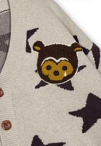 Cardigan con bottoni in beige chiaro con motivo a stelle marrone scuro. Presenta una toppa ricamata testurizzata di un orsetto marrone e giallo sul petto.