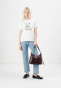 T-shirt blanc avec un motif d'ours en peluche, jean bleu clair et sac à main en cuir bordeaux avec une boucle en argent. Des mocassins noirs complètent le look.