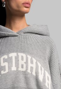 Sweat à capuche gris en tricot avec un motif texturé, arborant un logo blanc sur la poitrine et une capuche avec des bords côtelés.