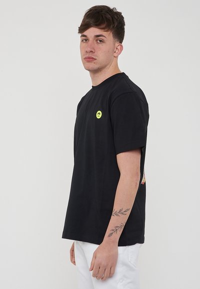 T-shirt en coton noir à manches courtes, col rond, avec un petit logo de smiley jaune à l'avant. Pantalons blancs.
