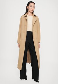Helle beige Trenchcoat mit einem doppelt geknöpften Design, Reverskragen und langen Ärmeln. Kombiniert mit schwarzen weiten Hosen. Glatte Textur.