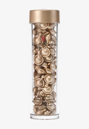 Contenant cylindrique transparent rempli de capsules de sérum en forme de goutte, de couleur or, étiquetées "Capsules de Céramide de Vitamine C." Un capuchon doré en haut.